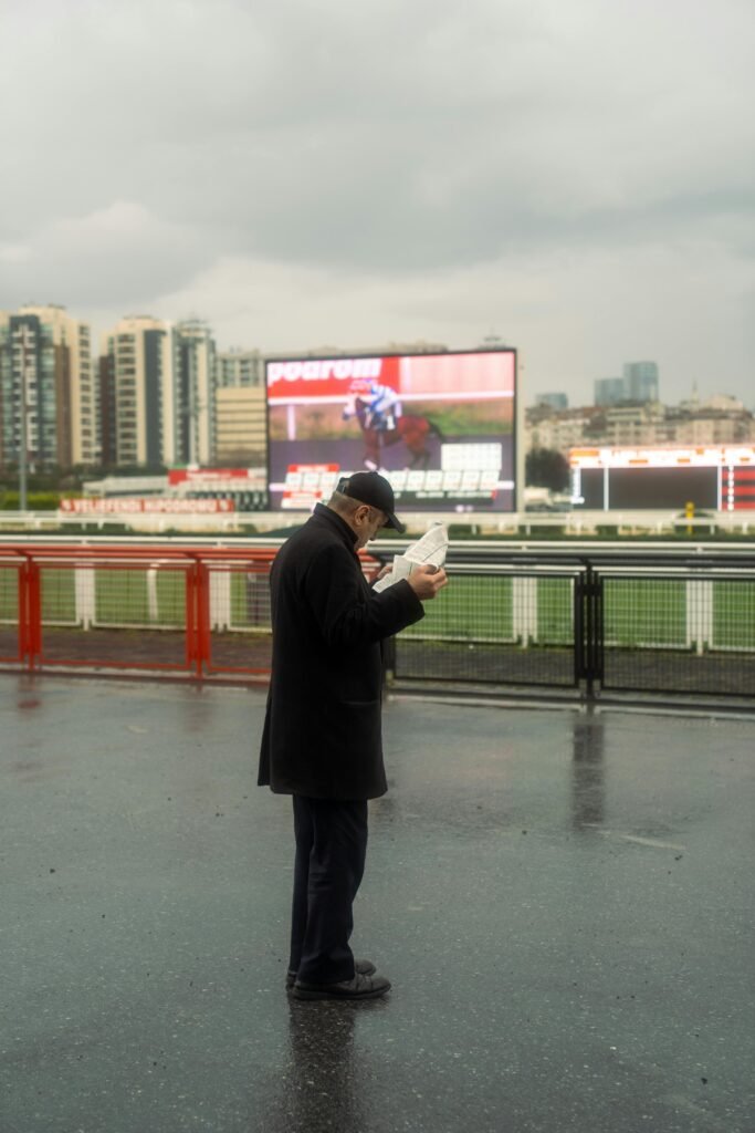 An elderly man reads a newspaper at a rainy horse race track with city skyline in the background.